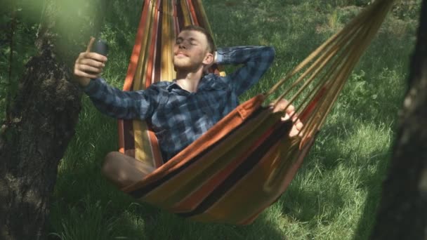 El hombre con el teléfono inteligente está relajando en la hamaca. Joven turista masculino tomando foto selfie en el teléfono móvil en hamaca al aire libre. El hombre toma selfies en smartphone en hamaca en jardín verde — Vídeo de stock