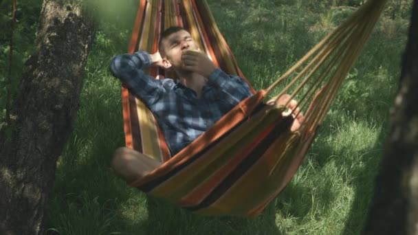 カジュアルな服装の男性フリーランサーは、本質的にハンモックに横たわっている。ハンモックの上で屋外でリラックス目を閉じた男。田舎で週末を楽しむ男性観光客。キャンプだ。夏休みだ。ハンモックでリラックス — ストック動画