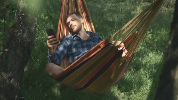 Surfer sur Internet mobile. Homme avec téléphone cellulaire dans les mains se détendre dans l'hamac à l'extérieur. Détendez-vous dans l'hamac. Jeune touriste masculin couché sur hamac et utilisant le téléphone mobile. Camping. Vacances d'été — Video