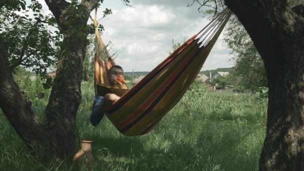 Hombre caucásico guapo que relaja en la hamaca y que lee el libro en el día soleado del verano al aire libre. Joven macho acostado en hamaca en el jardín verde. Turista relajante al aire libre en hamaca. Acampar. Vacaciones de verano — Vídeos de Stock