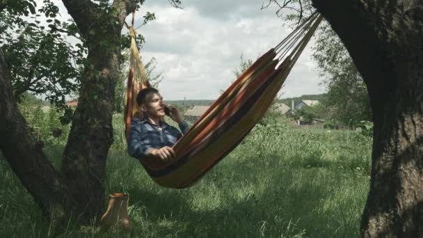 Hombre hablando por teléfono móvil en hamaca al aire libre. Hombre hablando por teléfono inteligente mientras está acostado en hamaca entre dos árboles en un lugar acogedor campo. Hombre disfrutando de vacaciones y columpiándose en hamaca — Vídeo de stock