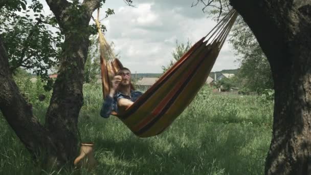 En glad man som ligger i hängmatta och blåser bubblor. Ung man blåser bubblor samtidigt koppla av i hängmatta på grön trädgård. Sommarsemester koncept. Man kopplar av i hängmatta utomhus. Slappna av i hängmattan — Stockvideo