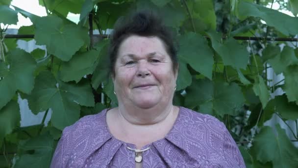 Mujer mayor sonríe a la cámara, retrato. Mujer vieja caucásica feliz en vestido de pie en el patio. Mujer de mediana edad sonriendo. Mujer mayor de mediana edad mirando a la cámara. Anciana jubilada sonriendo — Vídeo de stock