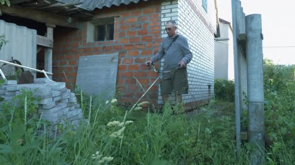 Facet kosi wysoką trawę na podwórku w letnim domu. Mężczyzna ogrodnik koszenie zielonej trawy z kosiarką wzdłuż podwórka. Facet używający kosiarki na zewnątrz. Kosiarka tnąca zieloną trawę — Wideo stockowe