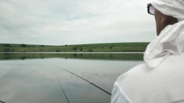 Balıkçı tekneyle gölde balık tutuyor. Nehir kıyısında elinde olta olan bir adam oturuyor. Erkek gölde balık yakalar. Arka planda güzel bir doğa vardır.