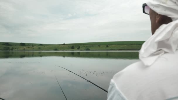 Ψαράς ψαρεύει ψάρια στη λίμνη με βάρκα. Άντρας με καλάμι ψαρέματος να κάθεται κοντά στην ακτή. Αρσενικά αλιεύματα ψαριών στη λίμνη με όμορφη φύση στο παρασκήνιο — Αρχείο Βίντεο