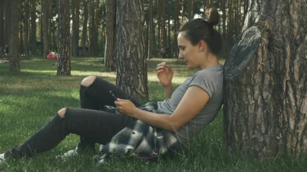 Vrouw zittend op haar skateboard op gras in het stadspark. jong duizendjarig meisje zitten buiten met skateboard en kijken naar het scherm op de telefoon in het park — Stockvideo