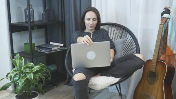 在笔记本电脑上工作的女孩坐在椅子上。在家工作。呆在家里年轻的女性在家里用笔记本电脑在现代公寓舒适的椅子上工作。妇女开始在总公司的笔记本上工作 — 图库视频影像