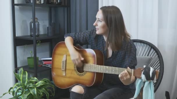 Vrouwelijke gitarist die thuis akoestische gitaar speelt. Vrouw speelt akkoorden op gitaar zitten in gezellige stoel in modern appartement. jong charmant meisje leren gitaar spelen — Stockvideo