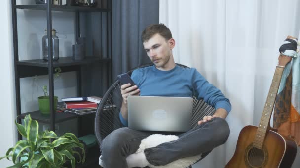 Hombre joven relajante en silla acogedora en el apartamento moderno, utilizando el teléfono y el ordenador portátil. Hombre sentado en la oficina en casa con ordenador portátil. Empresario con teléfono de chat y comunicación en línea en Internet — Vídeos de Stock