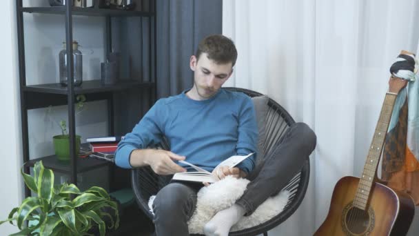 Joven sentado en una silla acogedora en la sala de estar moderna y libro de lectura. Hombre relajante y lectura libro en casa. Hombre en casa sentado en la silla delante de la ventana y libro de lectura — Vídeos de Stock