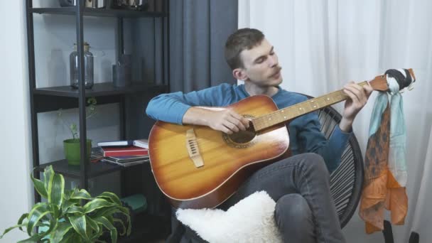 Uśmiechnięty, beztroski młodzieniec gra na gitarze akustycznej i śpiewa w domu w przytulnym salonie. mężczyzna muzyk gra na gitarze siedzi w krzesło w domu — Wideo stockowe