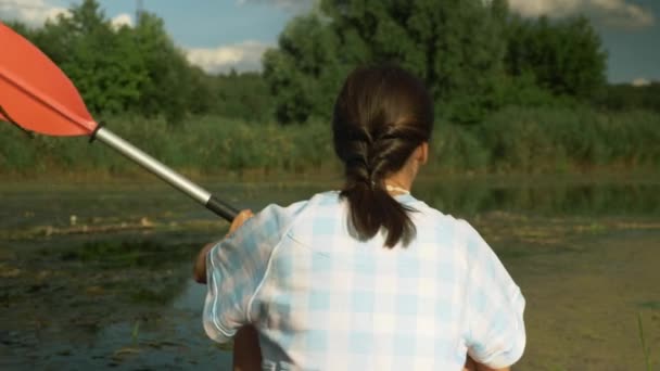 Sportowa kobieta fitness unosi się w kajaku na spokojnej rzece latem, strzał w plecy. Dziewczyna jest kajakarstwo i zwiedzanie rzeki podczas wakacji letnich. Widok z tyłu młodej samicy z wiosłem na łodzi na jeziorze — Wideo stockowe