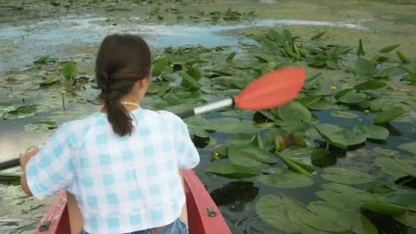 若いブルネットの女性はハスの花を通してカヌーで帆。女性旅行者は夏に川のカヤックに浮かんでいます。観光客は休日の週末に川でボートで美しい自然景観を探索します — ストック動画