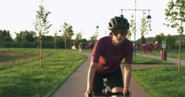 Genç bir kadın gün batımında yaz parkında bisiklet sürüyor. Mutlu kadın bisiklet yolunda bisiklet sürüyor. Güneş gözlüklü ve kasklı profesyonel bisikletçi, gün ışığında motosikletle pedal çeviriyor.