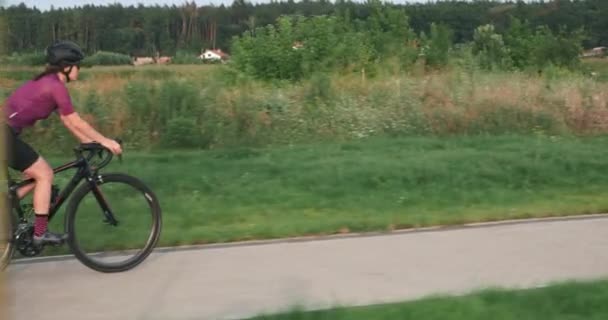 Athletic fit woman cyclist is cycling on road bike on promenade. Professional triathlete is training on bicycle and preparing for triathlon competition. Sports female is riding on cycle path at sunset — Stock Video