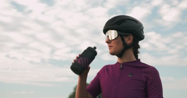 Porträtt av professionell cyklist dricksvatten från flaska mot blå himmel. Brunett kvinna i svart hjälm och cykling bära drycker isoton dryck efter träning på cykel. Triathlonkonceptet — Stockvideo