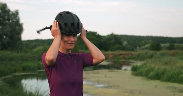 Портрет молодой женщины-велосипедистки в черном матовом шлеме, стоящей в парке на закате. Спортивная самка надевает шлем перед поездкой на велосипеде. Улыбающаяся девушка надевает шлем перед велогонкой — стоковое видео