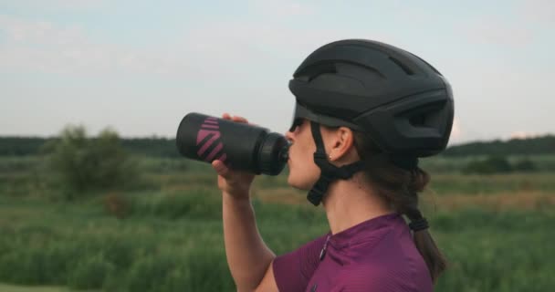 Уставшая счастливая велосипедистка в солнцезащитных очках и шлеме пьет воду после тяжелой тренировки на велосипеде на закате. Триатлонистка пьет воду из бутылки для гидратации во время тренировки по велоспорту — стоковое видео