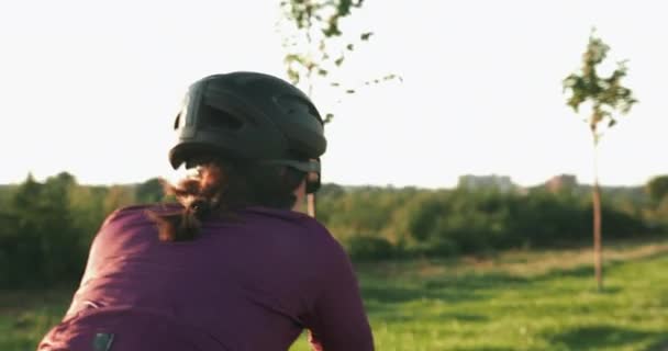 自転車での女性の後ろ姿は日没時に公園に前方に移動します。ヘルメットの若い女性サイクリストは、太陽の下でロード自転車に乗ってサイクリングしています。プロのトライアスリートは日の出に自転車でトレーニングしています。スポーツコンセプト — ストック動画