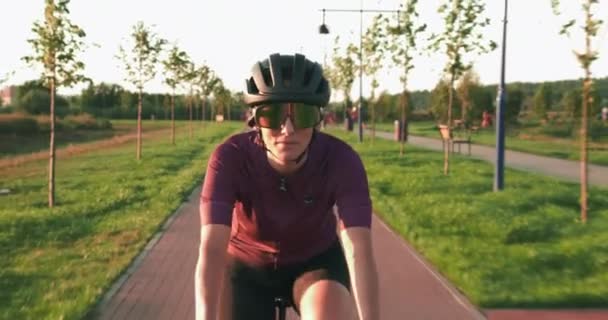 フィット運動サイクリストは、太陽の光の下で道路のエアロバイクにペダルを回転させている。若い女性アスリートの肖像画は、日没時に自転車道でペダルを漕いでいます。幸せなやる気のある女性が自転車に乗っている — ストック動画