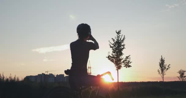 Radfahrerin steht mit Fahrrad auf Hügel und fotografiert am Telefon den schönen goldenen Sonnenuntergang. Mädchen genießt Sonnenuntergang nach Radtour. Sportlerin entspannt sich nach Radtraining bei Sonnenuntergang — Stockvideo