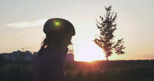 La silueta de la muchacha ciclista que habla por teléfono en los rayos del sol en el anochecer. Mujer joven en el casco está de pie cerca de la bicicleta y hablando por teléfono, sonriendo y disfrutando de la hermosa puesta de sol de oro. Concepto de viaje — Vídeo de stock