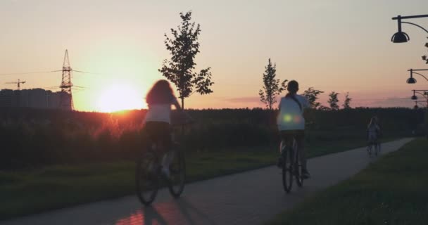 Ludzie jeżdżą rowerami po ścieżce rowerowej w parku o zachodzie słońca. Szczęśliwa rodzina spędza czas razem w parku w słońcu. Dzieci bawią się w parku latem o zachodzie słońca. Szczęśliwy styl życia — Wideo stockowe