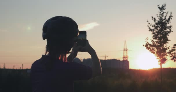 Chica está haciendo fotos de hermosa puesta de sol en el teléfono. Silueta de ciclista mujer está tomando fotos en el teléfono contra la luz del sol. Mujer viajera de bicicleta de pie junto a la bicicleta contra el atardecer dorado — Vídeo de stock