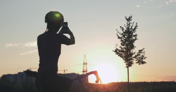 Una viaggiatrice sta scattando foto al tramonto. Donna con bici è in piedi nel parco contro la bella luce del sole e facendo foto sul telefono. Concetto di viaggio. Silhouette di ragazza in casco — Video Stock