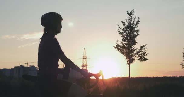 La silueta de la chica en el casco es beber agua después del paseo en bicicleta. Mujer joven bebe agua después de aventuras en bicicleta al atardecer. Viajero de bicicletas está de pie en el fondo de los edificios durante el atardecer de oro — Vídeo de stock