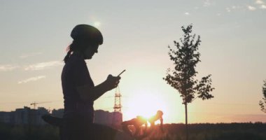 Kadın gün batımında parkta dinleniyor. Genç bayan sporcu bisikletin yanında duruyor, doğanın ve güneşin tadını çıkarıyor. Altın gün batımında şehir binalarının arka planında duran bisikletçi silueti