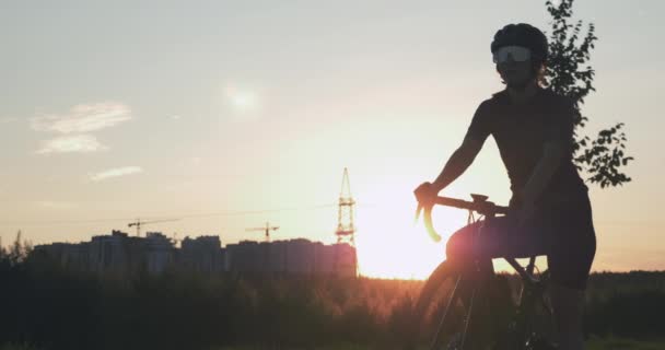 Silhuett av ung kvinna cyklist mot solnedgången. Kvinnlig idrottsman står i parken i solsken och börjar rida på cykel. Passform flicka börjar cykla i parken vid soluppgången. Triathlonkonceptet — Stockvideo