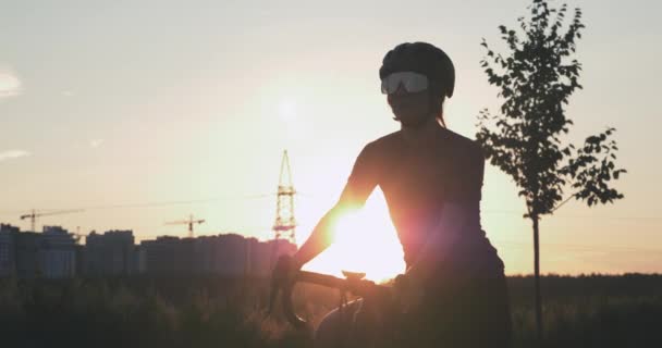 若い女性は美しい夕日に対して公園に立っており、自転車のペダルをねじれ始めています。太陽の光の下で魅力的な女性サイクリストの肖像画。プロのトライアスリートが自転車でペダルを踏む — ストック動画