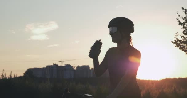 若い美しい女性の肖像画は日没時に水を飲む。女性サイクリスト公園でのハードトレーニングの後に水を飲んでいる太陽の光がいっぱい。サイクリングとトライアスロンのコンセプト。健康的なライフスタイル — ストック動画