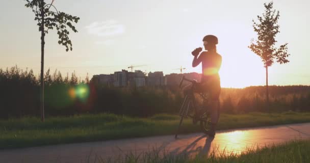 Femme cycliste boit de l'eau après une balade à vélo dans le parc contre le coucher du soleil. Portrait de la jeune athlète boit de l'eau après l'entraînement cycliste, debout sur la piste cyclable sous les rayons du soleil. Concept de triathlon — Video