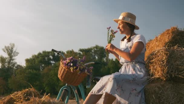Charming young girl is resting on meadow at sunset, her blue vintage retro bike parked next to her. Woman with old bicycle on field in sunshine. Portrait of pretty woman in stylish outfit at sunset — Stock Video