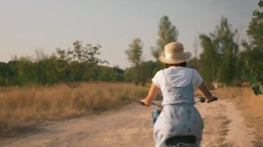 Büyüleyici romantik kız gezgin güneş ışığında kırsal yolda retro motorla pedal çeviriyor. Gün batımında bisikletli mutlu bir kadın. Yazlık elbiseli ve hasır şapkalı genç bir kadın tarlada bisiklet sürüyor.