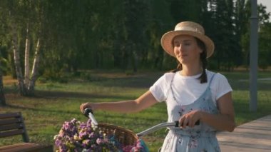Sundress 'li genç romantik gezgin kadın gün batımında şehir parkında klasik bisikleti yürütür ve yuvarlar. # Kız temiz havada yürüyor # # geçmişe dönük bir bisikletle # # Çiçeklerle dolu sepetle güneş ışığında #