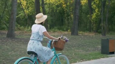 Şapkalı ve yazlık elbiseli genç kız elinde çiçeklerle klasik bir bisiklet sürüyor ve gülümsüyor. Kadın bisikletçi gün batımında parkta eski bir bisikletle bisiklet sürüyor. Güzel bayan bisikletçi parkta eğleniyor.