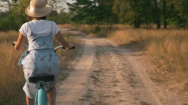 Girl is cycling on vintage bicycle through meadow at sunset. Woman In hat and sundress with bike on holiday riding in park. Beautiful pretty young brunette female rides bicycle on sunset filled park — Stock Video