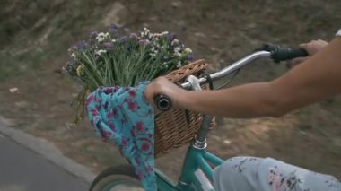 Retro bisiklet gidonunu çiçekli saman sepetle kapat. Kız parkta bisiklet sürüyor. Yazlık elbiseli kadın güneşli bir günde şık bir bisiklete biniyor. Mutlu kadın tatilde eğleniyor..