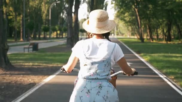 Okouzlující dáma v klobouku a modré šaty je jízda na kole s květinami na slunci. Mladá žena cyklistka je na kole v parku při západu slunce. Bruneta dívka jezdí retro kolo na cyklistické silnici v městském parku — Stock video
