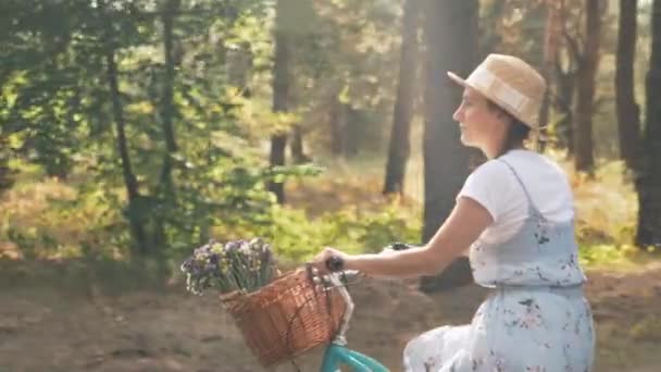 Beztroska urocza dziewczyna w kapeluszu i retro stylowa sukienka pedałuje na niebieskim rowerze z kwiatami w koszyku. Radosna rowerzystka jeździ na rowerze retro w słoneczny dzień w parku. Kobieta dobrze się bawi. — Wideo stockowe