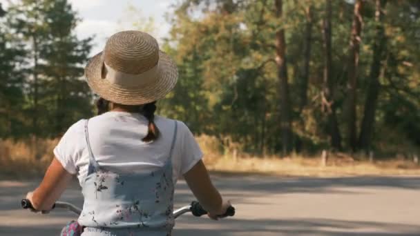 Chica feliz alegre es el ciclismo en bicicleta en el parque soleado en verano. Mujer morena paseos vintage bicicleta retro en el bosque con luz solar. Dama de sombrero y vestido se divierte y disfruta de las vacaciones de verano — Vídeo de stock