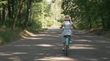 Esmer çekici kadın güneşli yeşil parkta bisiklet sürüyor. Güzel genç kız yazın ormanda antika bisiklet sürüyor. Mavi elbiseli ve şapkalı bayan eski moda bisiklet sürüyor.