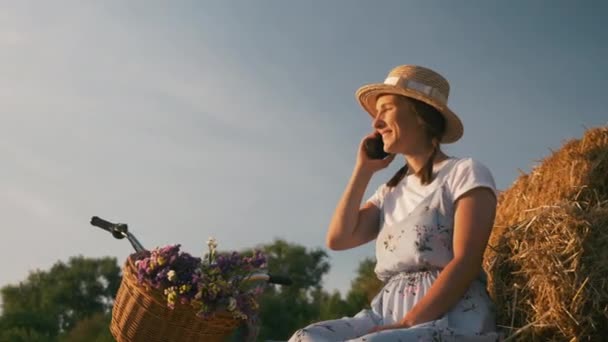 Femme cycliste en tenue rétro élégante parle par téléphone au coucher du soleil après avoir roulé à vélo. Femme est assise sur la paille près du vélo rétro vintage, souriant et parlant par téléphone le soir d'été au soleil — Video