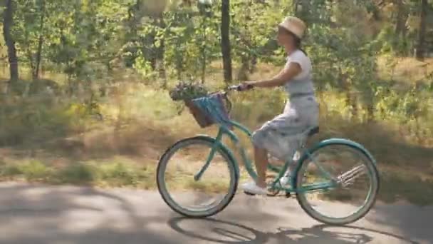 Ευτυχισμένη γυναίκα βόλτες vintage ποδήλατο με λουλούδια στο καλάθι, απολαμβάνοντας ηλιόλουστη μέρα στο πάρκο. Ξέγνοιαστη μελαχρινή κοπέλα σε ψάθινο καπέλο και sundress σε ρετρό ποδήλατο το καλοκαίρι ηλιόλουστο δάσος στον ήλιο. Στυλ ρετρό — Αρχείο Βίντεο