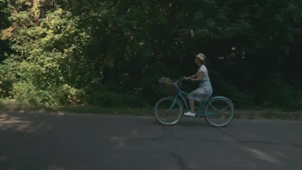 Usmívající se žena cyklistka je na kole ve slunečném parku v létě. Šťastná mladá dívka se pohybuje na retro kole v městském parku. Žena v klobouku a šaty na staromódní kolo v městském parku za slunečného dne — Stock video