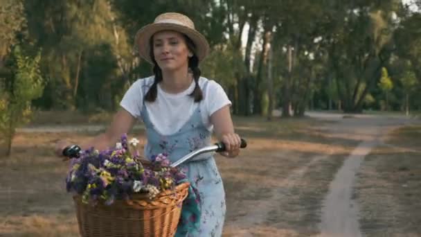 Portrait de jeune femme charmante aux yeux romantiques rêveurs en robe de soleil et chapeau sur vélo rétro vintage à la lumière du soleil. Happy fille souriante est à vélo le long de la prairie au coucher du soleil — Video